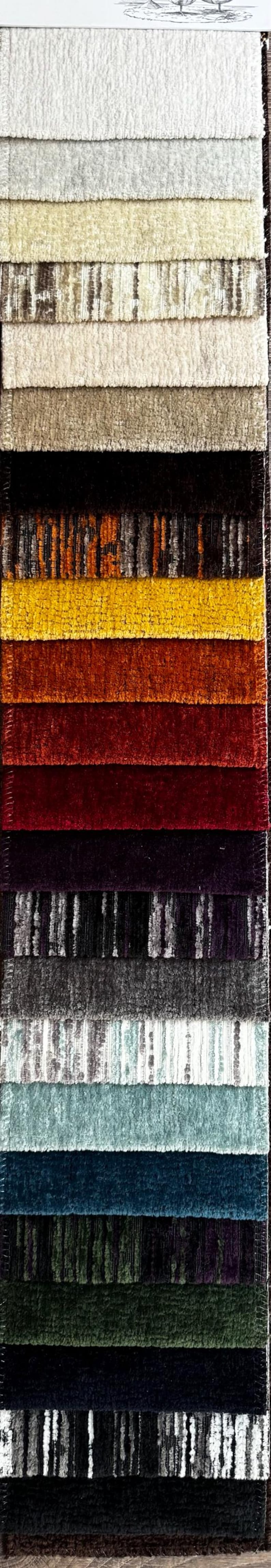 May include: A vertical display of fabric swatches in various colors and textures. The samples range from light neutral tones to deep jewel tones, including shades of beige, brown, gold, red, purple, and teal. The fabrics appear to be plush and textured.