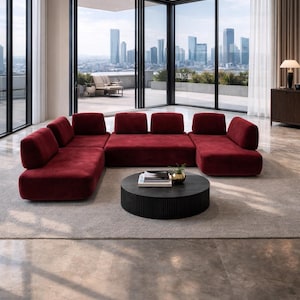 May include: A modern living room featuring a large, modular sectional sofa in a deep red hue. A round, black coffee table sits in front of the sofa, and large windows offer a city view. The room has a neutral color palette.
