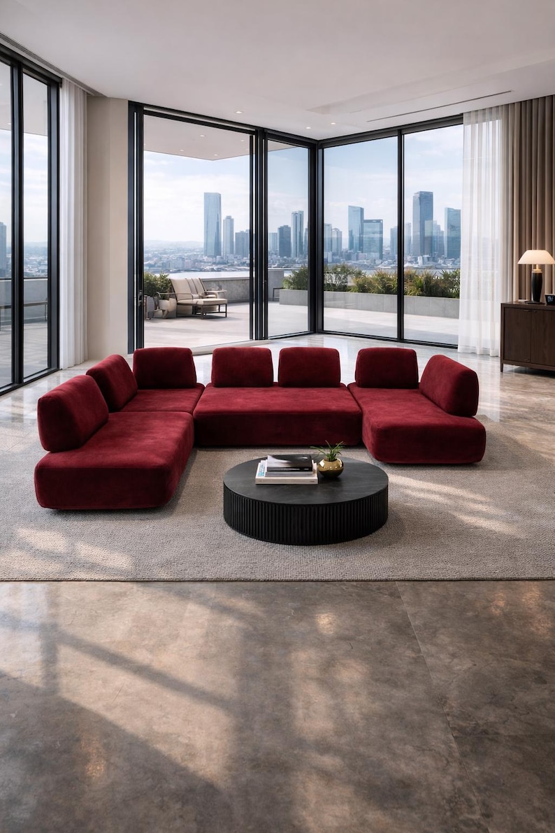 May include: A modern living room featuring a large, modular sectional sofa in a deep red hue. A round, black coffee table sits in front of the sofa, and large windows offer a city view. The room has a neutral color palette.