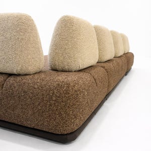 May include: A modern sofa with a brown textured base and light tan, rounded back cushions. The sofa has a low profile and a dark brown border along the base. The cushions are arranged in a row.