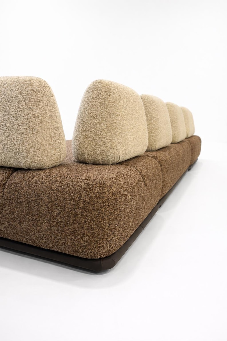 May include: A modern sofa with a brown textured base and light tan, rounded back cushions. The sofa has a low profile and a dark brown border along the base. The cushions are arranged in a row.