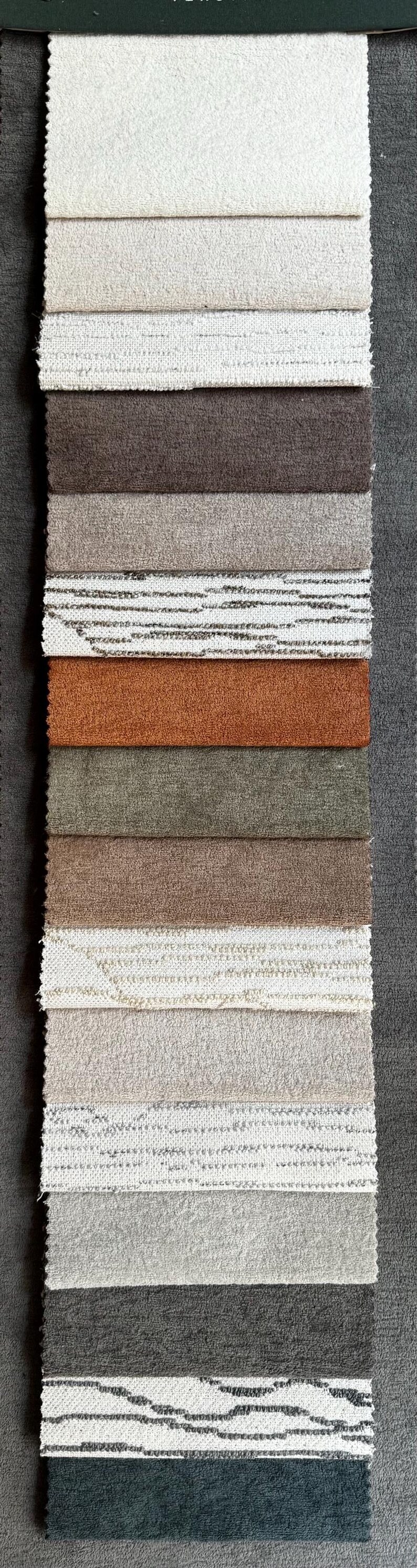 May include: A vertical display of fabric swatches in various neutral colors, including white, beige, brown, and gray. The textures vary from smooth to textured, with some featuring patterned designs. Ideal for upholstery or home decor.