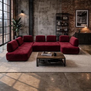 May include: A modern living room featuring a large, modular burgundy sofa. A dark wood coffee table sits on a neutral rug. The room has exposed brick and concrete walls, with a large window and a black metal bookshelf.