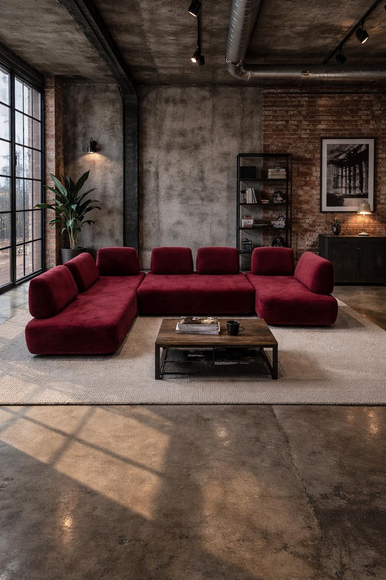 May include: A modern living room featuring a large, modular burgundy sofa. A dark wood coffee table sits on a neutral rug. The room has exposed brick and concrete walls, with a large window and a black metal bookshelf.