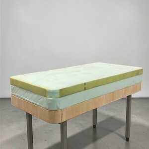 May include: A rectangular table with a light green mattress top and a light wood base. The table has four silver metal legs. The mattress appears to be made of foam. The table is set against a white background.