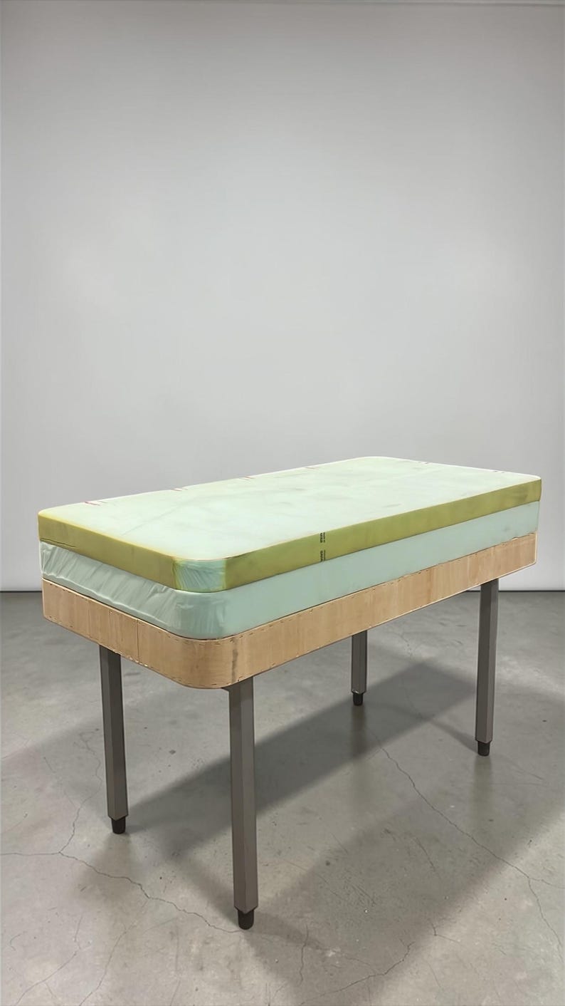 May include: A rectangular table with a light green mattress top and a light wood base. The table has four silver metal legs. The mattress appears to be made of foam. The table is set against a white background.