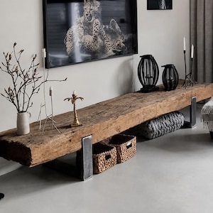 Puede incluir: Un banco largo y rectangular hecho de una viga de madera gruesa y desgastada con una superficie rugosa y texturizada. El banco está sostenido por dos patas de metal robustas en forma de U. Está decorado con un jarrón blanco y cestas.