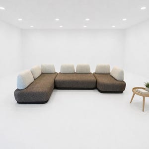 May include: A modern sectional sofa with a dark brown base and light-colored cushions. A small, round wooden side table with a plant and books sits nearby. The room is minimalist with white walls and a white floor.