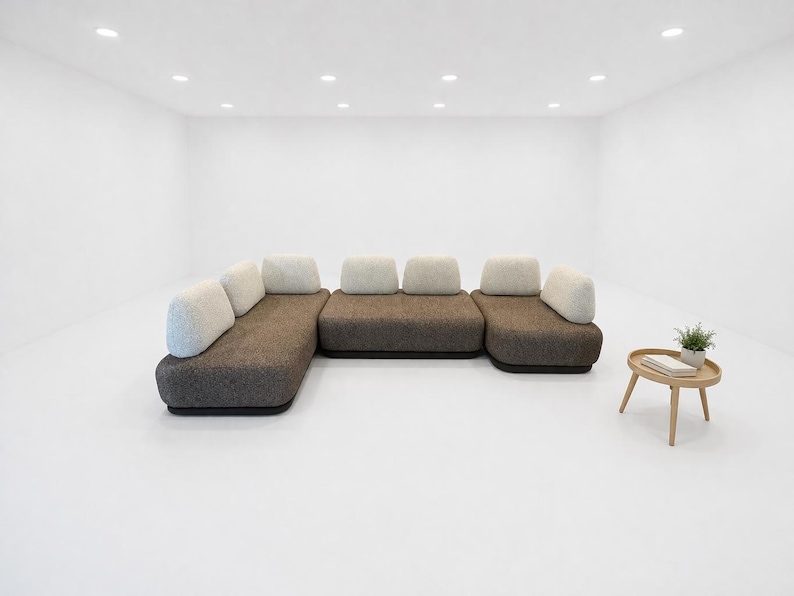 May include: A modern sectional sofa with a dark brown base and light-colored cushions. A small, round wooden side table with a plant and books sits nearby. The room is minimalist with white walls and a white floor.