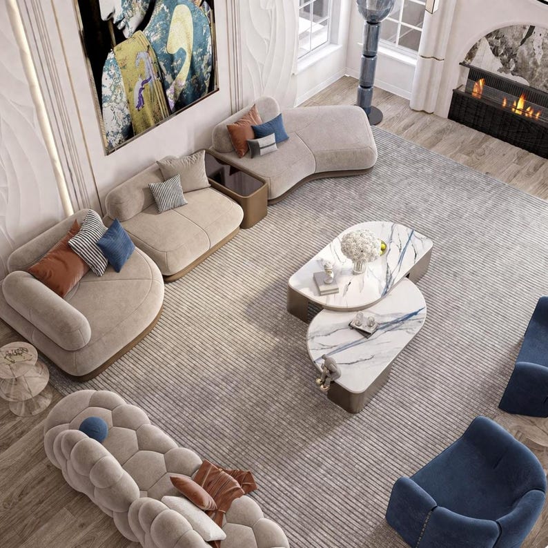 May include: An overhead view of a modern living room with beige curved sofas, blue armchairs, and a patterned rug. Two marble-topped coffee tables are in the center, and decorative pillows add color. A fireplace and artwork complete the stylish interior.