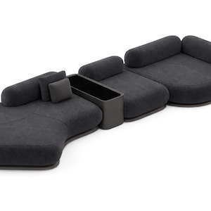 May include: Modular dark gray sofa with rounded shapes and a built-in storage unit. The sofa has several sections, including a curved chaise lounge, a loveseat, and a single seat, all with soft, textured upholstery. Includes two cylindrical and one square pillow.
