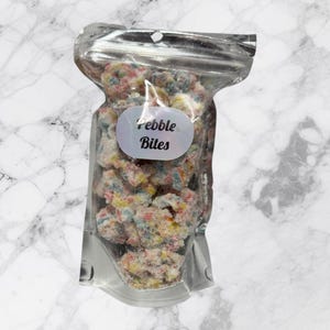 Może przedstawiać: Srebrna, zamykana torebka wypełniona kolorowymi kęsami płatków zbożowych. Torebka ma etykietę z napisem "Pebble Bites".