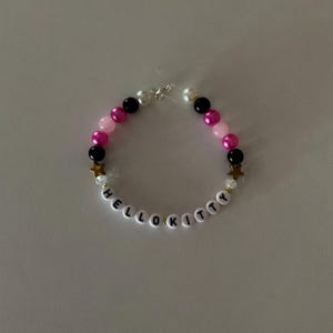 May include: A Hello Kitty beaded bracelet with pink, black and white beads, gold star charms, and white letter beads spelling out "Hello Kitty".