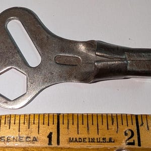 Könnte beinhalten: Ein metallischer Aufziehschlüssel mit einem flachen, unregelmäßigen Griff, der einen sechseckigen Ausschnitt und einen länglichen Schlitz aufweist. Der Schlüssel liegt über einem Holzlineal, das eine Gesamtlänge von etwa 5,7 Zentimetern zeigt.