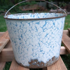 May include: A round metal bucket featuring a white and blue marbled enamel finish. The bucket has a thin, rusted metal handle attached to the top rim. The bottom edge shows significant brown rust and wear, with a matching rusted metal rim.