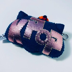 K&ouml;nnte beinhalten: Ein marineblauer Filz-Schl&uuml;sselanh&auml;nger mit dem Wort "POP" in metallisch ros&eacute;goldenen Buchstaben. Die Buchstaben sind mit gr&uuml;nem Faden gen&auml;ht. Ein kleiner orangefarbener Akzent ist oben sichtbar. Ein silberner Verschluss ist angebracht.