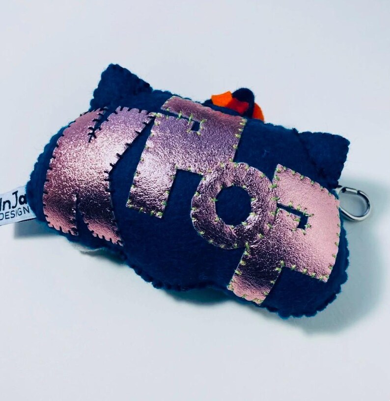 K&ouml;nnte beinhalten: Ein marineblauer Filz-Schl&uuml;sselanh&auml;nger mit dem Wort "POP" in metallisch ros&eacute;goldenen Buchstaben. Die Buchstaben sind mit gr&uuml;nem Faden gen&auml;ht. Ein kleiner orangefarbener Akzent ist oben sichtbar. Ein silberner Verschluss ist angebracht.