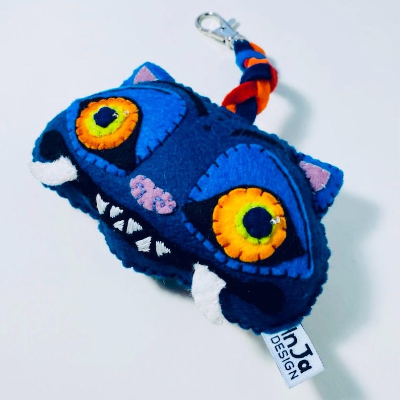 K&ouml;nnte beinhalten: Ein handgefertigter blauer Filz-Schl&uuml;sselanh&auml;nger in Form eines Monsterkopfes. Das Monster hat gro&szlig;e orange und gelbe Augen, wei&szlig;e Z&auml;hne und rosa Akzente. Ein geflochtener orangefarbener, blauer und roter Riemen ist mit einem silbernen Verschluss verbunden. Das Etikett lautet "InJa DESIGN".