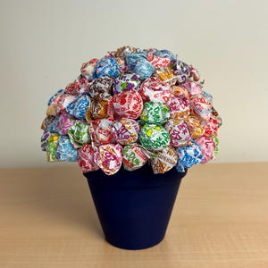 Puede incluir: Una maceta de cerámica azul llena de piruletas envueltas de colores, creando un arreglo floral único y festivo.