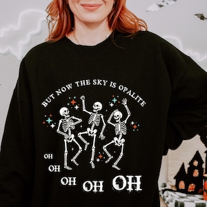 Pode incluir: Camisola preta com um desenho de três esqueletos a dançar e o texto "BUT NOW THE SKY IS OPALITE" e "OH OH OH". O design inclui estrelas e detalhes coloridos, ideal para o Halloween.