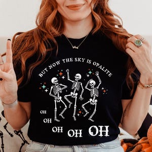 Könnte beinhalten: Schwarzes T-Shirt mit einer Grafik von drei tanzenden Skeletten und dem Text "BUT NOW THE SKY IS OPALITE" in Weiß. Das Design enthält bunte Sterne und das Wort "OH" wiederholt. Das T-Shirt wird von einer Person getragen.