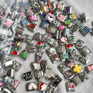 May include: A collection of stainless steel charm bracelets with various colorful designs. Charms include a soccer ball, a teddy bear, a pink star, a red poppy, a house, and a flower. Some charms have text, including "MOM" and "The Beatles".