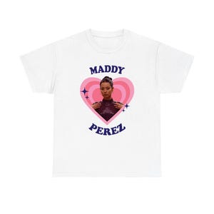Pode incluir: Camiseta branca com um gráfico de uma mulher dentro de um coração rosa, com o texto "Maddy Perez" em azul. O design inclui estrelas rosa. A camisa é feita de um material macio e confortável.