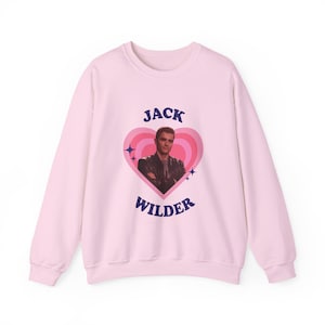 Könnte beinhalten: Hellrosa Sweatshirt mit herzförmigem Grafikdesign. Das Design zeigt ein Foto eines Mannes, rosa und weiße konzentrische Herzen, blaue Sterne und den Text "JACK WILDER" in Marineblau.