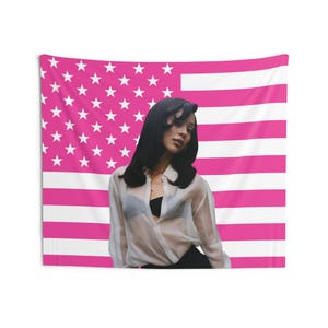 Può includere: Un arazzo con la bandiera americana rosa e bianca con una donna con una camicia bianca trasparente, un top nero e pantaloni neri. La donna ha lunghi capelli neri e indossa una collana d'oro.