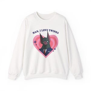 May include: White sweatshirt with a graphic of a bat-like creature in a suit inside a pink heart. The text "MAN, I LOVE TWINKS" is above the heart. The design includes blue star accents.
