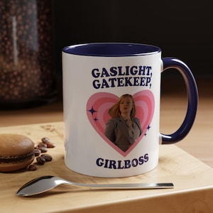 Peut inclure: Mug en céramique blanche avec intérieur et anse bleu marine. Le mug affiche le texte "GASLIGHT, GATEKEEP, GIRLBOSS" avec un cœur rose et une image de femme. Une cuillère et une pâtisserie sont au premier plan.