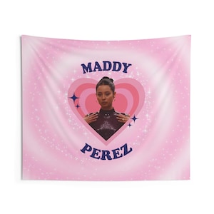 Può includere: Arazzo rosa con un design a forma di cuore con l'immagine di una donna. Il testo "MADDY" è sopra l'immagine e "PEREZ" sotto, entrambi in blu scuro. Lo sfondo ha un effetto sfumato con brillantini bianchi.
