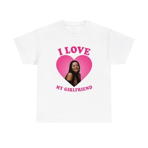 Può includere: T-shirt bianca con grafica a cuore rosa. Il cuore contiene una foto di una persona sorridente. Le parole "I LOVE MY GIRLFRIEND" sono stampate sopra e sotto il cuore in rosa.