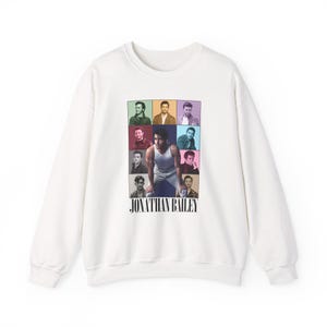 May include: White crewneck sweatshirt featuring a grid of colorful portraits and a central image of a man in a tank top. The text "JONATHAN BAILEY" is printed in black below the images. The sweatshirt is made of a soft material.