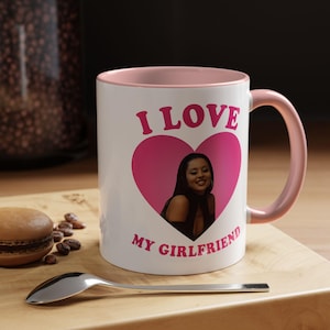 Può includere: Tazza in ceramica bianca con interno e manico rosa. La tazza presenta la scritta "I LOVE MY GIRLFRIEND" in rosa, con un cuore rosa contenente la foto di una donna. Un cucchiaio e un macaron sono in primo piano.