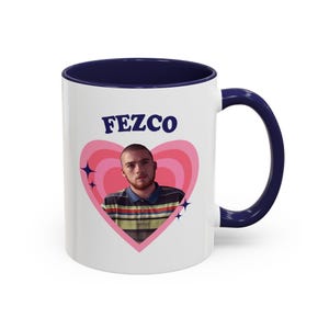 Può includere: Tazza in ceramica bianca con interno e manico blu navy. La tazza presenta un design a cuore rosa con un ritratto di una persona e la scritta "FEZCO" in blu navy. La tazza ha una capacità di 325 ml.