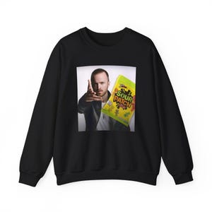 Peut inclure: Sweat-shirt noir avec un imprimé d'un homme pointant du doigt, tenant un paquet de bonbons Sour Patch Kids. Le sac est jaune, vert et rouge. Le sweat-shirt est de style décontracté.