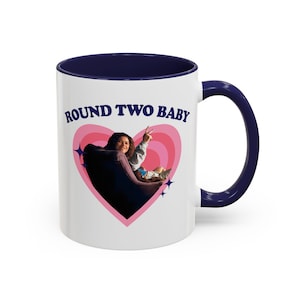 Può includere: Tazza in ceramica bianca con interno e manico blu navy. La tazza presenta la scritta "ROUND TWO BABY" sopra una grafica a cuore rosa con una foto di una persona all'interno. La tazza ha una finitura lucida.