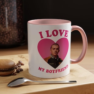 Può includere: Tazza in ceramica bianca con interno e manico rosa. La tazza presenta un cuore rosa con una foto di una persona all'interno e le parole "I LOVE MY BOYFRIEND" in lettere rosa. Un cucchiaio e un macaron sono in primo piano.