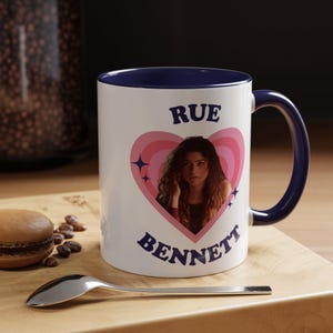 Può includere: Tazza in ceramica bianca con interno e manico blu. La tazza presenta un design a forma di cuore con la foto di una persona e la scritta "RUE BENNETT". Un cucchiaio e un pasticcino sono su una superficie di legno.