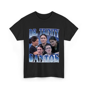 Può includere: T-shirt nero con una grafica di più donne e la scritta "DR. TRINITY" sopra e "SANTOS" sotto in un font blu retrò. Il design ha un'estetica vintage.