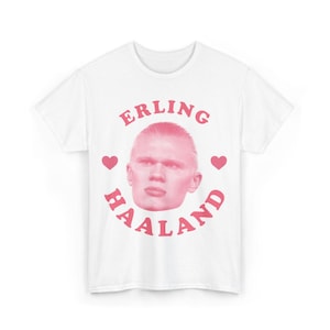 Peut inclure: Un t-shirt blanc à manches courtes avec une impression graphique rose sur le devant. Le motif comprend un portrait d'Erling Haaland centré dans une disposition circulaire du texte ERLING HAALAND et deux petits cœurs roses sur les côtés.