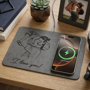 May include: A gray wireless charging pad with a sketch of a person holding a child, and the text "I love you!". A smartphone is charging on the pad. A framed photo, a succulent, and a cup of coffee are also on the wooden surface.