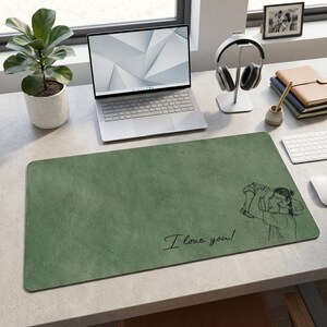 May include: Green desk mat with a textured surface. Features a black line drawing of a person holding a child, with the text "I love you!" in cursive. A laptop, headphones, and a small potted plant are also visible.
