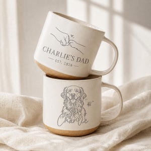 May include: Two stacked ceramic mugs with a speckled cream finish and a natural tan base. The top mug features a line drawing of a fist bump with a paw, the text CHARLIE'S DAD, and EST. 2026. The bottom mug shows a line drawing of a dog and the name Charlie.