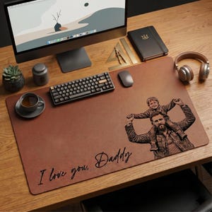 Op de afbeelding: Een bruine leren bureauonderlegger met de gegraveerde afbeelding van een man die een kind op zijn schouders draagt. De tekst "I love you, Daddy" is erop geschreven. Een toetsenbord, muis en andere kantoorbenodigdheden liggen op het houten bureau.