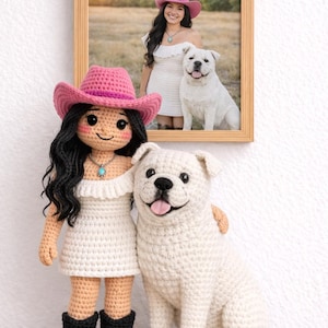 Puede incluir: Una muñeca de ganchillo con sombrero de vaquero rosa, vestido blanco y botas negras, está de pie junto a un perro blanco de ganchillo. Una foto enmarcada de una mujer con sombrero de vaquero rosa y un perro blanco cuelga en el fondo. La muñeca tiene pelo negro y un collar turquesa.