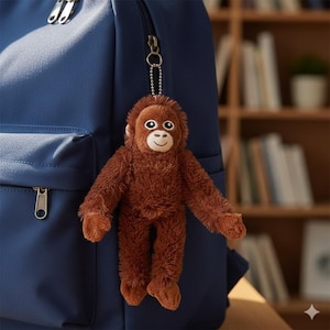 May include: A brown plush orangutan keychain with a smiling face and outstretched arms hangs from a navy blue backpack. The keychain is attached by a silver chain. The background includes a bookshelf.