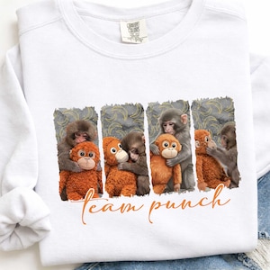May include: White sweatshirt with a graphic of monkeys hugging stuffed orangutans. The design is in a rectangular pattern with a gray background and gold banana shapes. The words "team punch" are written in orange script.