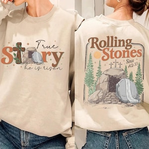May include: Beige sweatshirt with front and back graphics. The front features a cross, tomb, and text that reads "True Story, he is risen." The back has a "Rolling Stones" design with a tomb, trees, and text "Since AD 33."